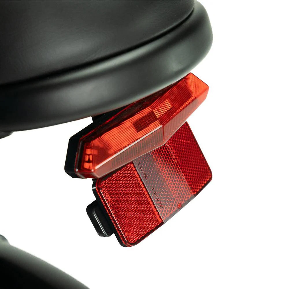 Close-up van een rode achterreflector en een lampje die onder een zwart fietszadel zijn bevestigd tegen een witte achtergrond, waarmee het stijlvolle ontwerp van de OUXI FATBIKE V8 PRO (C80 PRO) – MODEL 2026 met hydraulische remmen en alarmsysteem goed tot zijn recht komt.