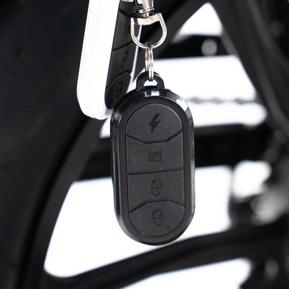 Een close-up van een zwarte elektronische sleutelhanger met drie knoppen die aan een metalen sleutelring hangt, bevestigd aan het frame van een OUXI FATBIKE V8 (C80) – MODEL 2026 met hydraulische remmen en een alarm.