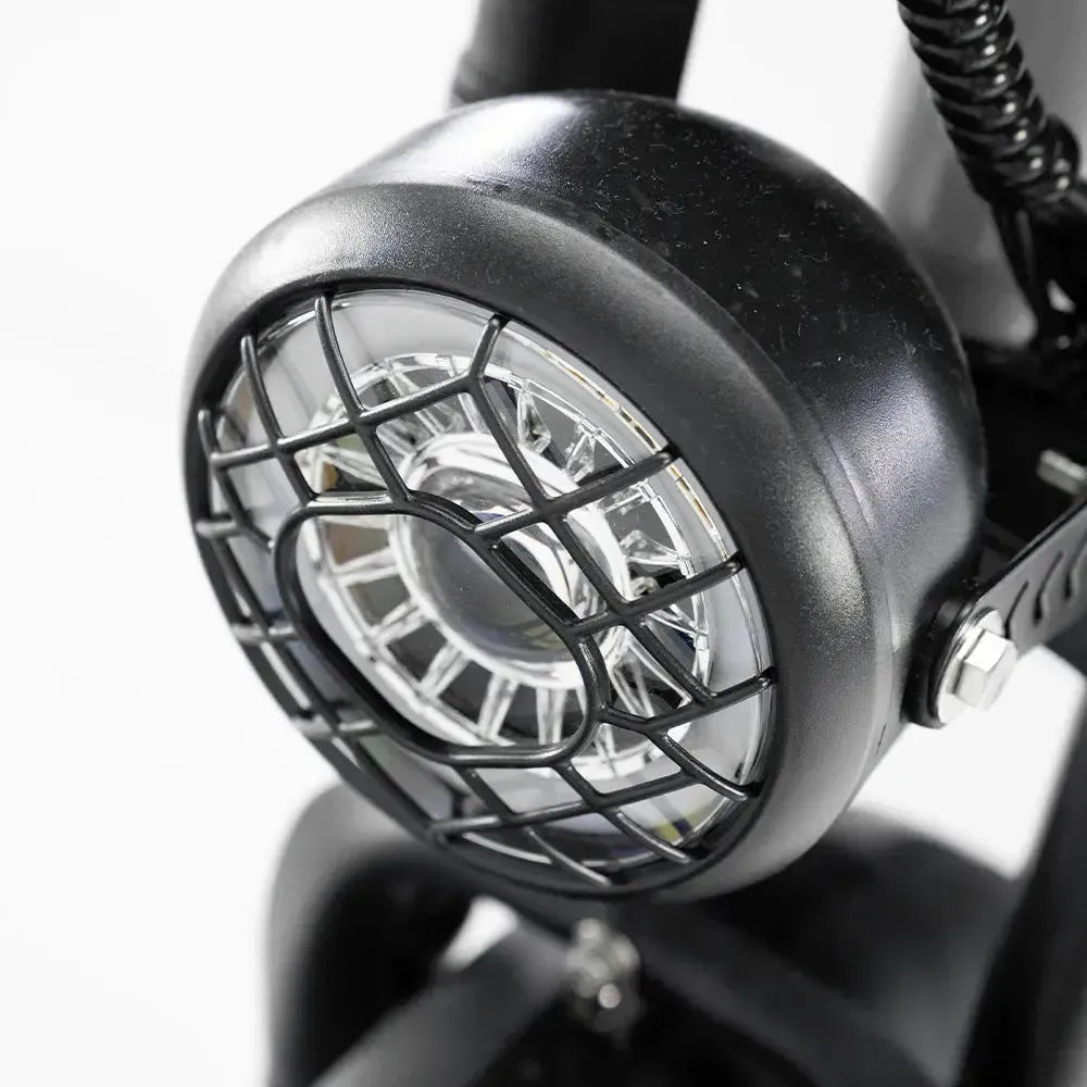 Close-up van een ronde koplamp met een beschermend zwart metalen rooster, gemonteerd op het zwarte frame van de OUXI FATBIKE OUXI C80 MINI – LICHT BLAUW, MODEL 2026, een elektrische fatbike met een 250W-motor, hydraulische remmen en een alarm, afgebeeld tegen een witte achtergrond.