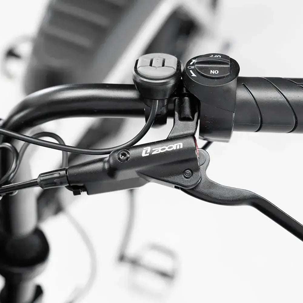 Close-up van een zwart stuur met een versnellingshendel, een remhendel met het opschrift „Zoom“ en diverse bedieningskabels, tegen een wazige wit-grijze achtergrond — waarmee de geavanceerde FATBIKE OUXI C80 MINI wordt getoond - ZWART (MODEL 2026) met hydraulische remmen en een alarmsysteem van OUXI.