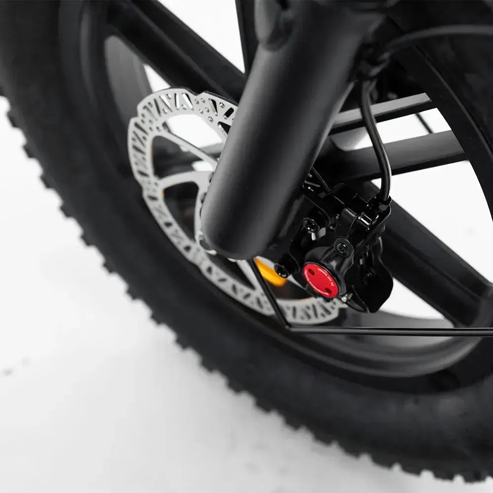 Close-up van het voorwiel van de OUXI FATBIKE C80 MINI - ZWART – MODEL 2026, met het hydraulische schijfremsysteem met een zwarte remklauw en rode accenten, gemonteerd op een zwarte vork, allemaal afgebeeld tegen een witte achtergrond – een strakke en krachtige elektrische fatbike van OUXI, ideaal voor gebruik in de stad.